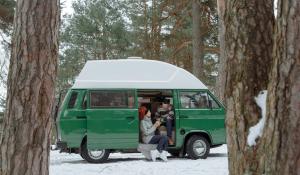a couple sitting in their green van that they live out of. It is a snowy day and they are wearing sweaters and holding warm mugs, smiling at each other.